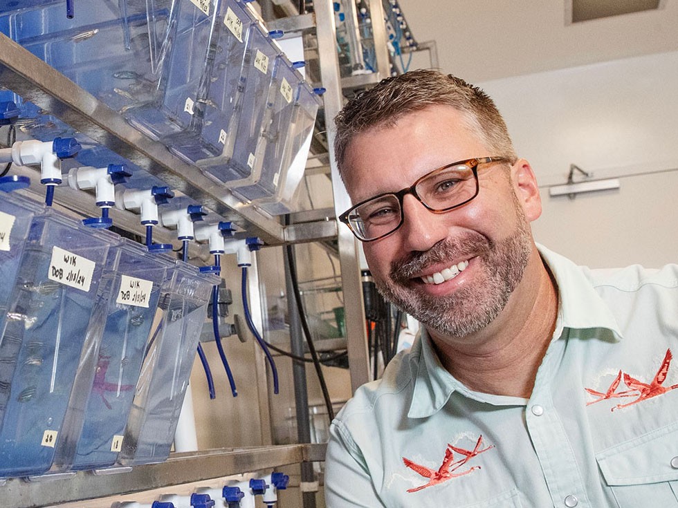 Biology professor, Jason Breves, smiles in a research lab with blue tanks on the left.