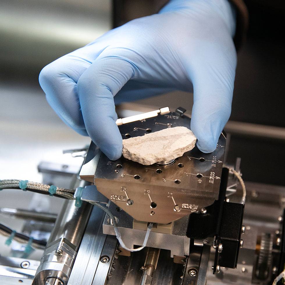 A hand wearing blue gloves places a piece of material on a microscopes platform 
