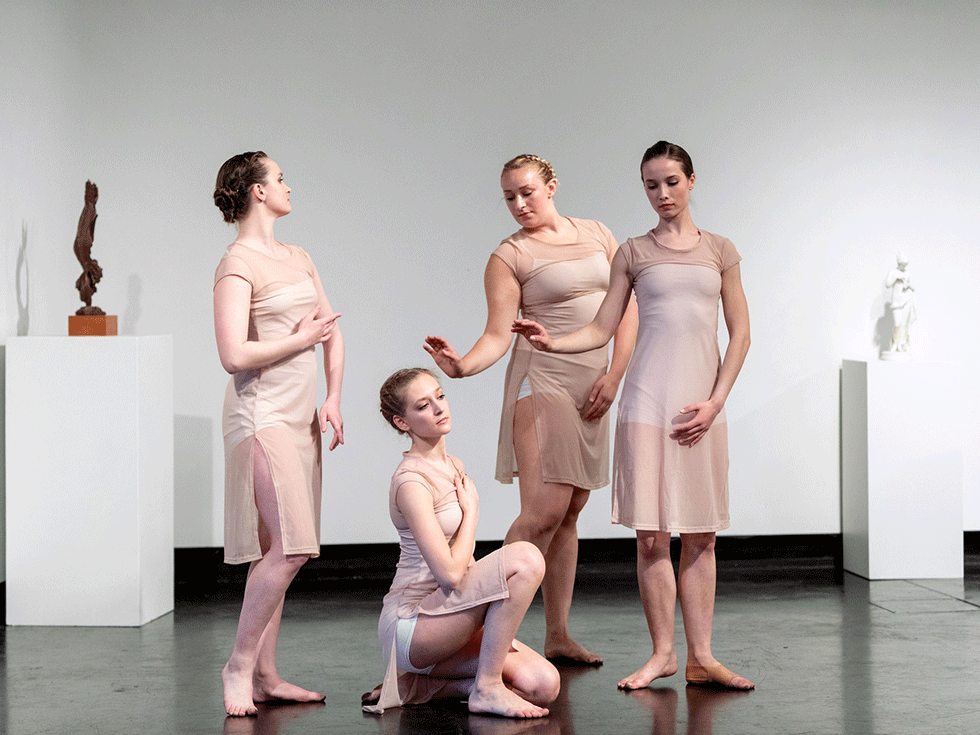 Four student dances perform in a gallery space at the Tang Museum. They are wearing pale pink flowy dresses, and each holds a different pose.