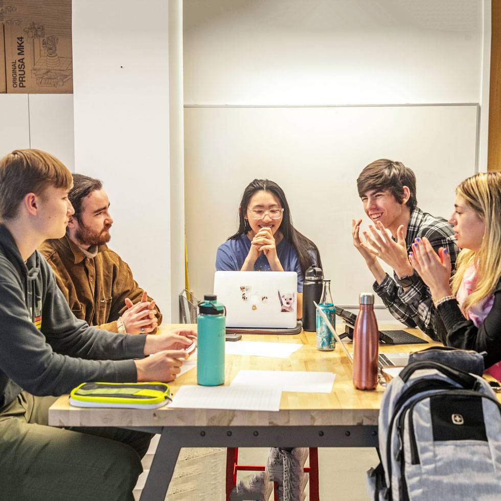 Five students sit around a table with their laptops, joyfully engaging in a classroom discussion.