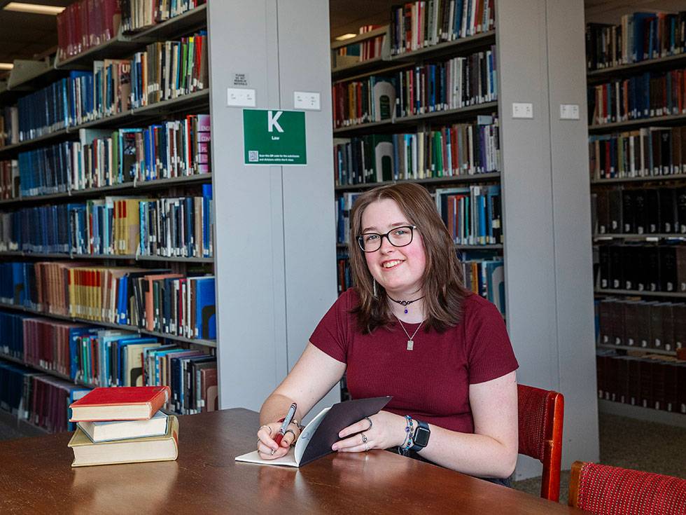 Skidmore College student Mia McCormack, a Greek scholar and peer tutor, works at a desk in the Lucy Scribner Library with book stacks in the background.