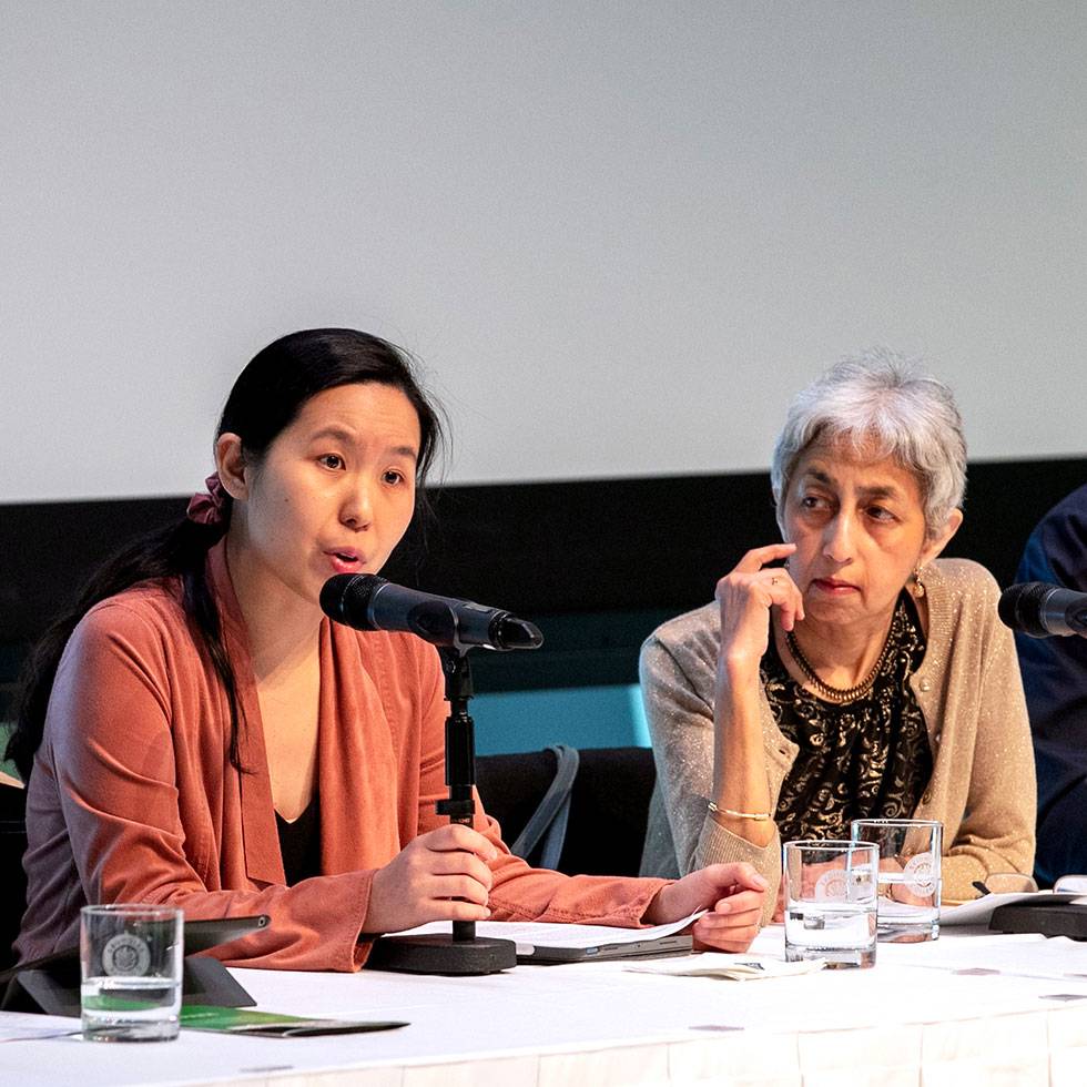 Jenny Day speaks during a panel discussion sitting at a table holding a microphone. Pushkala Prasad is seated to the right of the speaker.