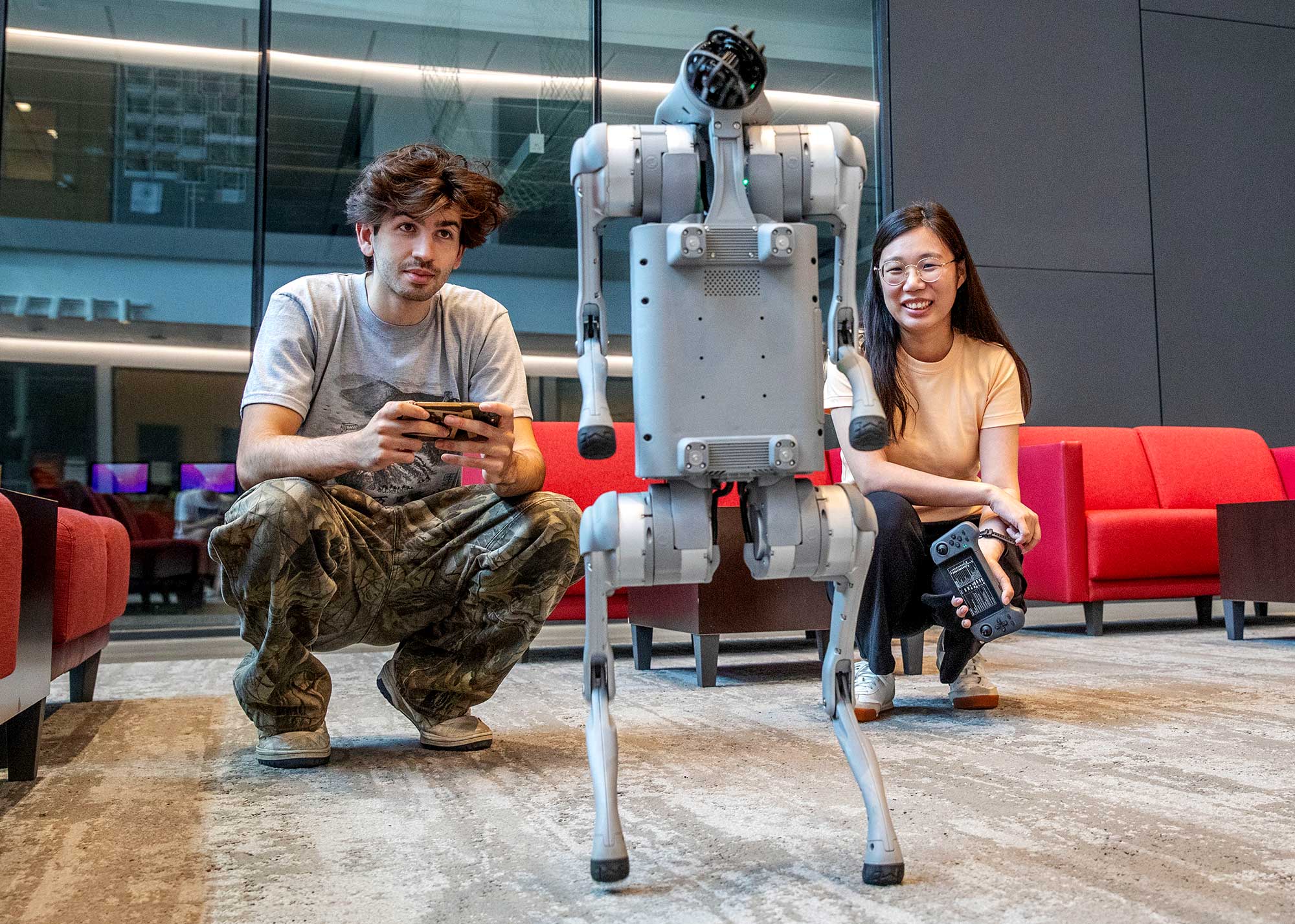 A student and a professor crouch indoors and smiling while operating handheld controllers as a robotic dog stands between them in a modern campus space.