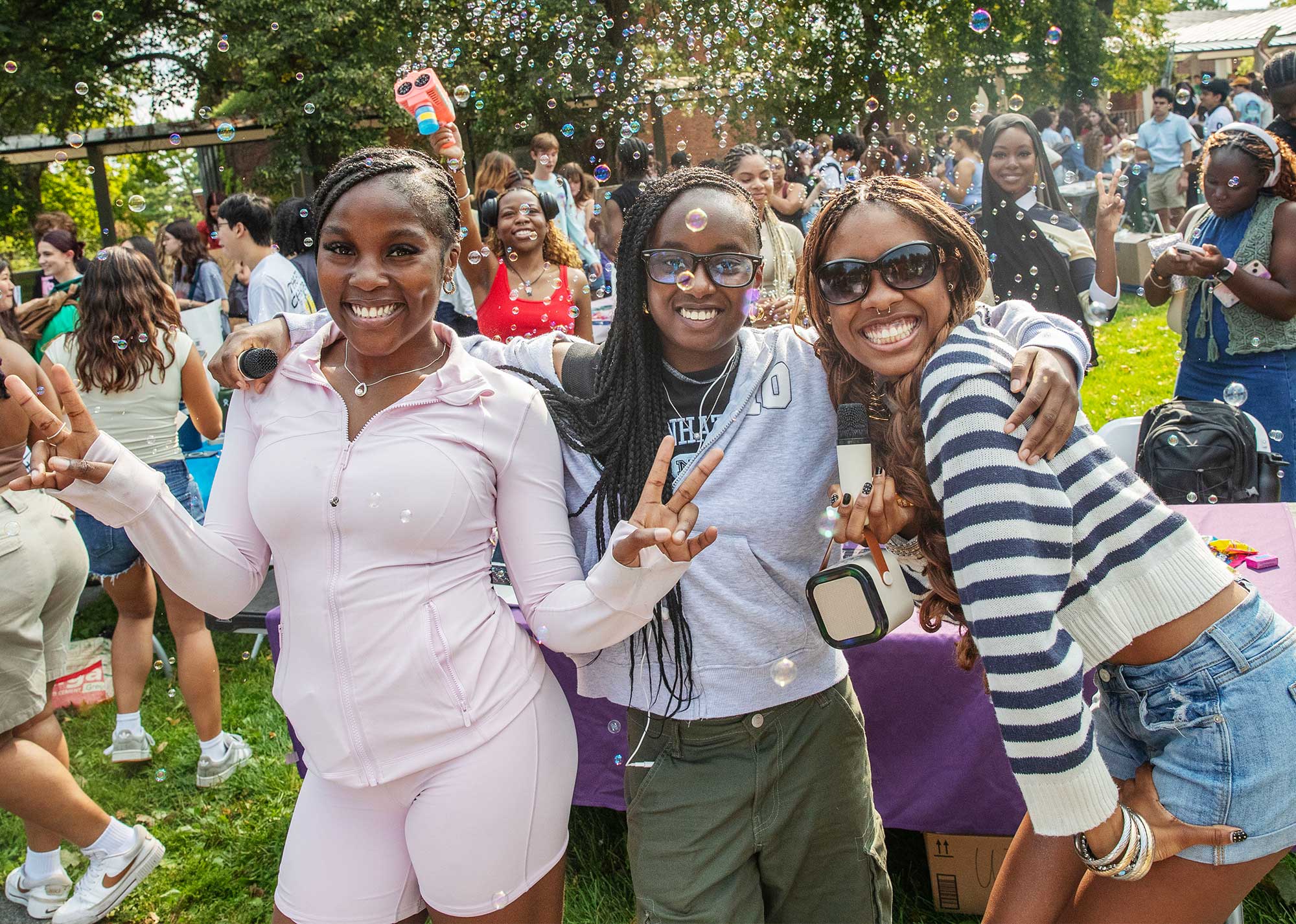 Three students smiling and posing together outdoors at a lively campus event, making peace signs as bubbles float around them and a crowd gathers in the background.