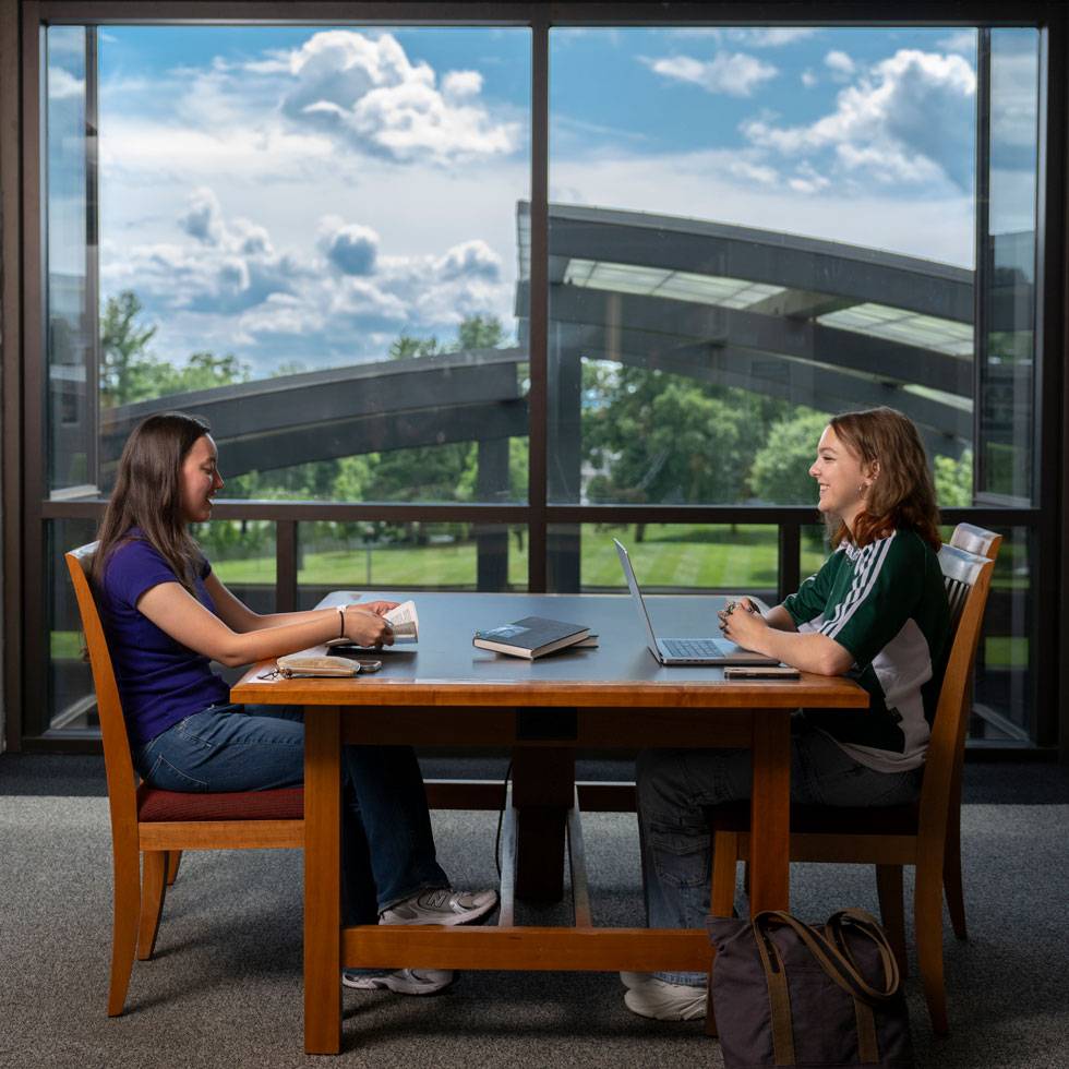 Two students smile at each other while sitting at a desk in the library overlook south park lawn