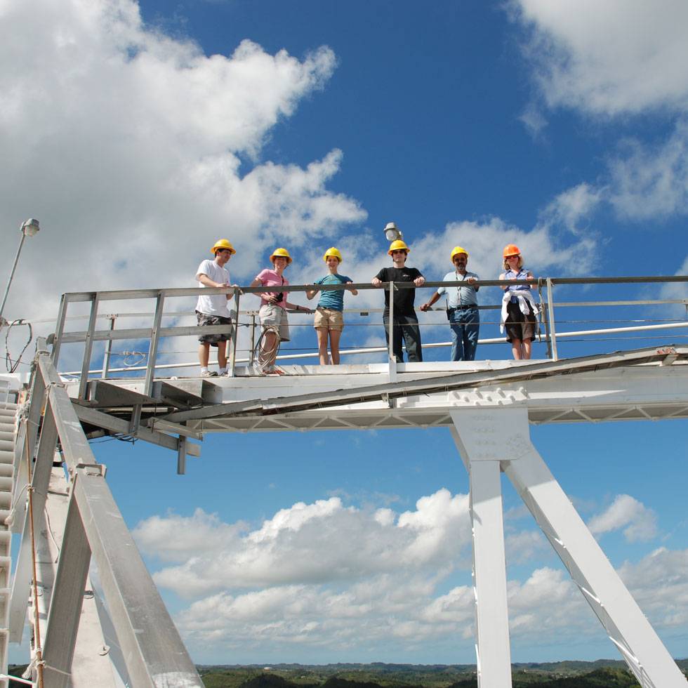 A group of people stand on a metal bridge with a blue sky in the background while doing research on an NSF grant