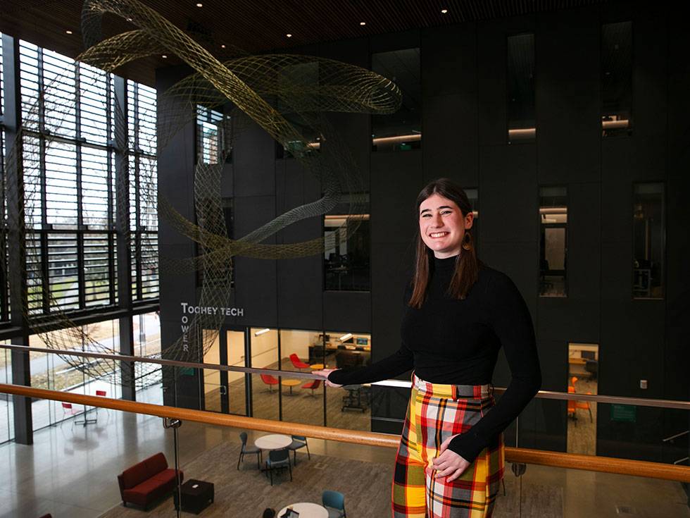 Student stands smiling on upper level balcony in Skidmore's Billie Tisch Center for Integrated Sciences, with the atrium and Toohey Tech Tower in the background