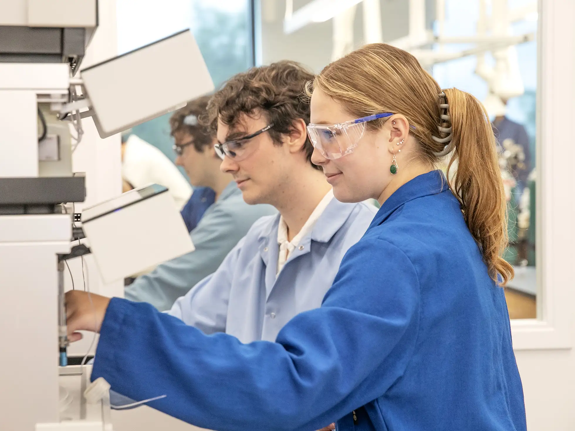 Two students wearing safety goggles and blue lab coats work together in a science lab, focusing on a large piece of analytical equipment.