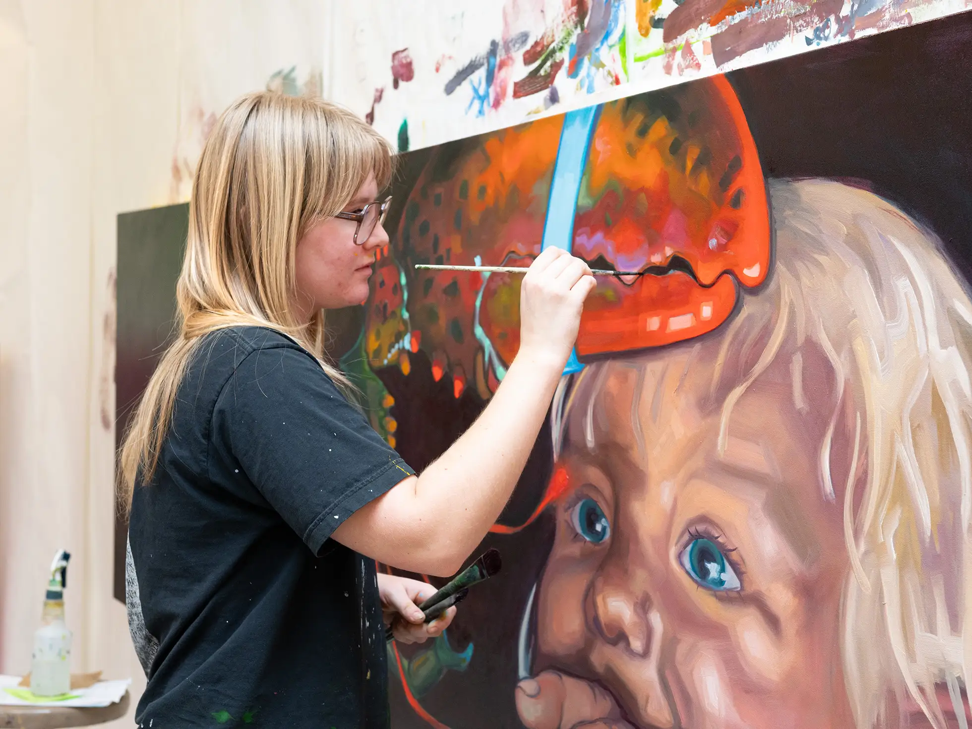 A Skidmore College student works on a large, colorful oil painting featuring a child's face and a lobster claw, showcasing fine arts studio work.