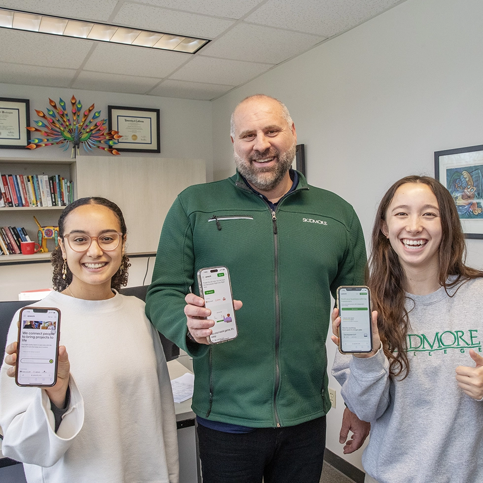 Two college students and a professor smile at the camera and show off an app on their phones that they created