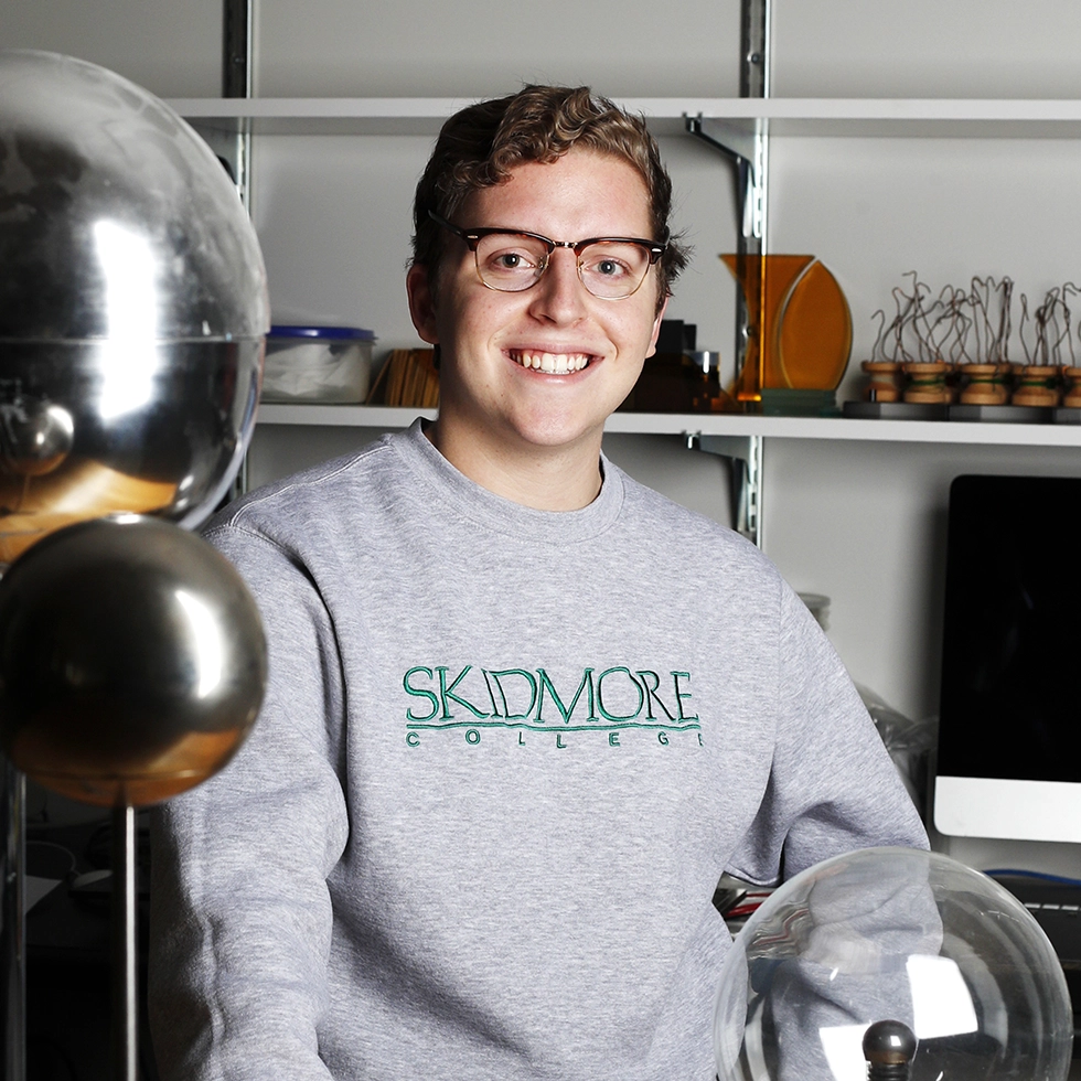 A young man in a gray sweatshirt that says Skidmore College poses in a physics laboratory for a portrait.