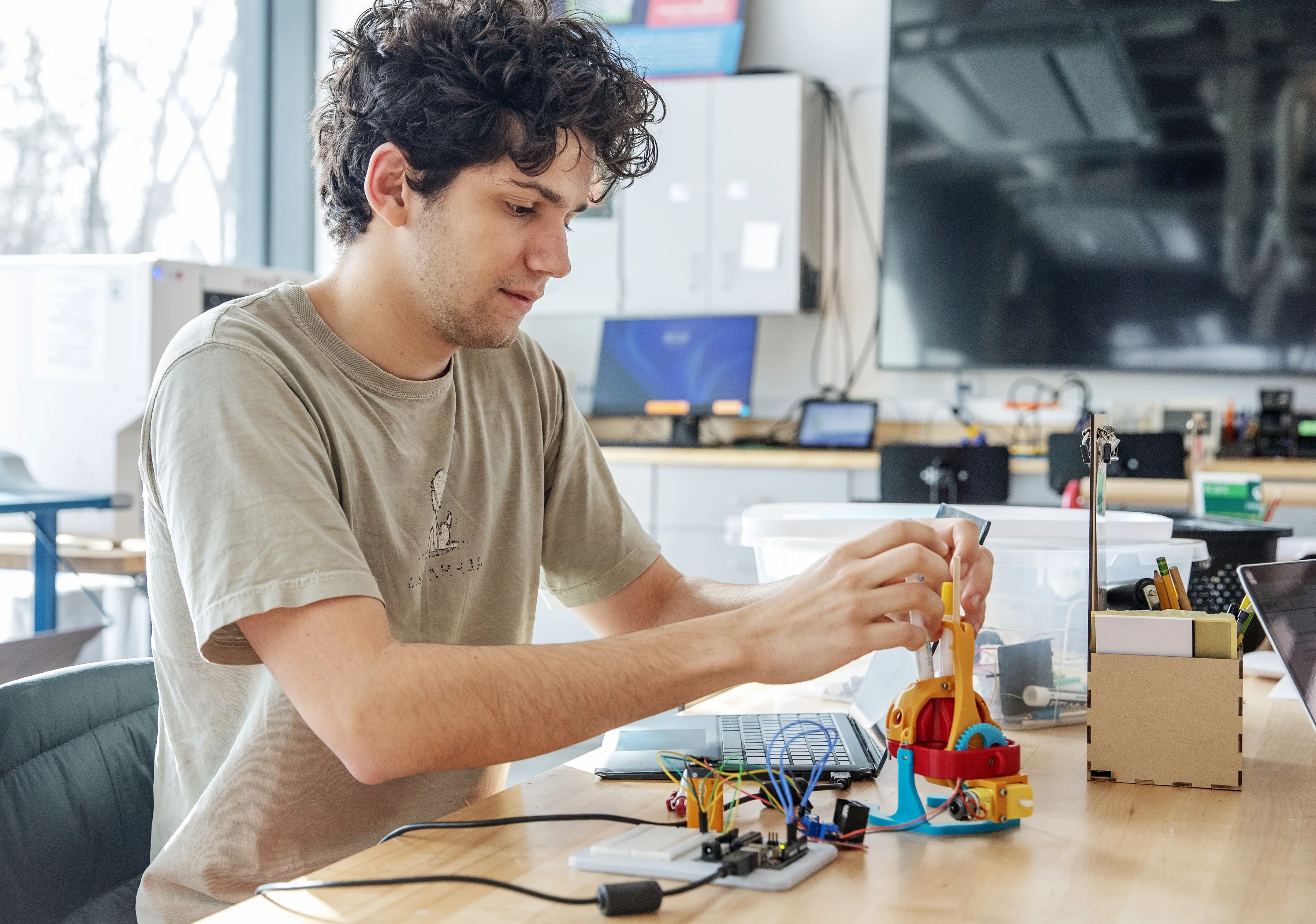A student concentrates on assembling a colorful mechanical device connected to circuits and wires at a workbench in a makerspace or innovation lab, surrounded by tools, laptops, and electronic components.