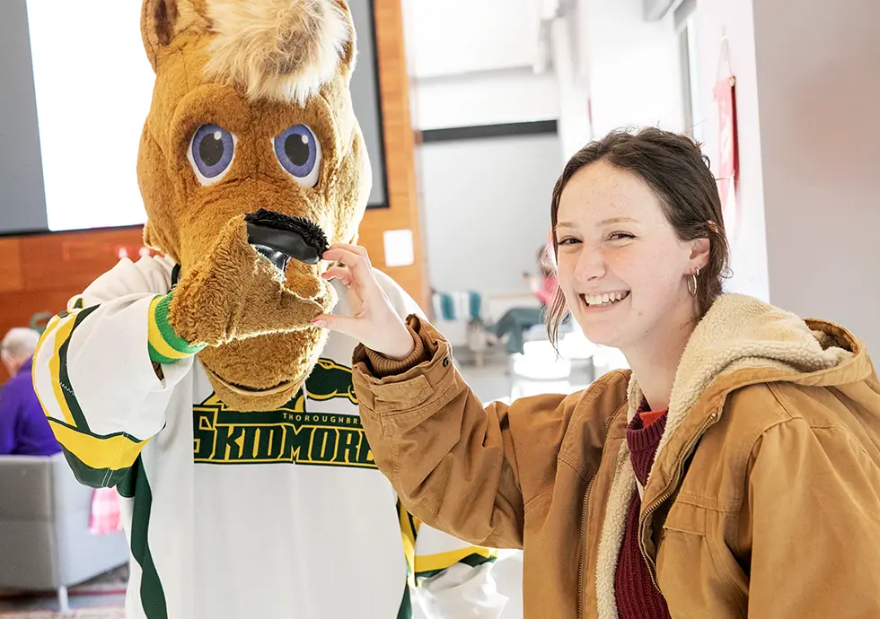 A smiling student makes a heart shape with their hands together with Skidmore College’s horse mascot, who is wearing a Skidmore Thoroughbreds jersey.