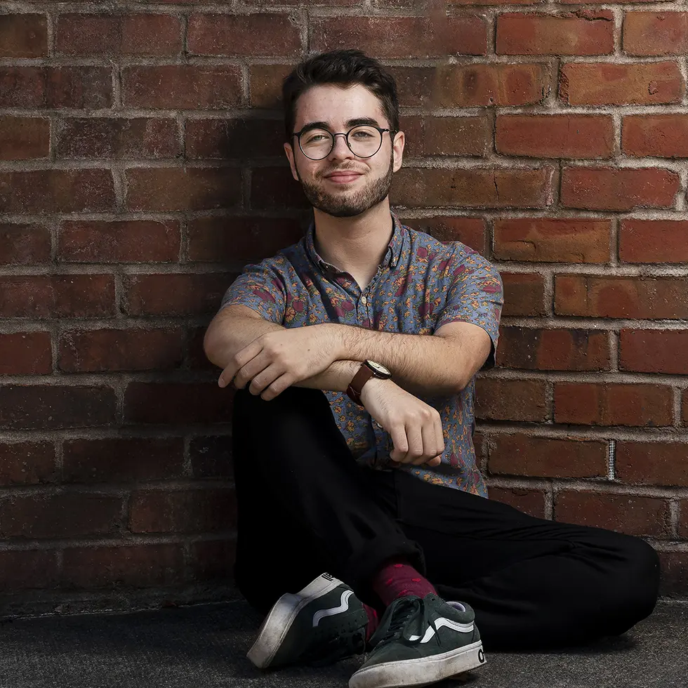 A student with glasses and a floral shirt sits on the ground, leaning against a brick wall with a casual and confident smile. His posture is relaxed, arms resting on his knees.