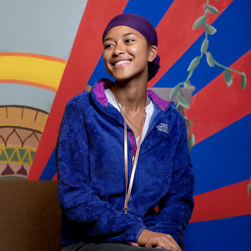 A student smiles brightly while seated in front of a colorful, mural-painted wall. She wears a blue North Face fleece and a purple headwrap, radiating warmth and positivity.