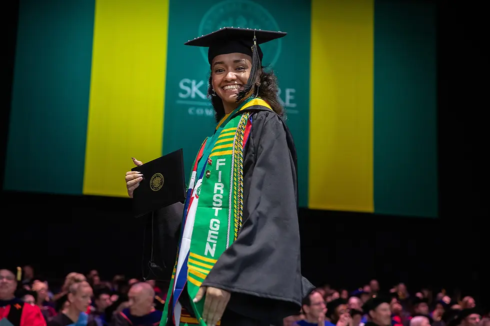 A smiling graduate on stage holds her diploma while wearing a cap, gown, and a green and gold “FIRST GEN” stole.