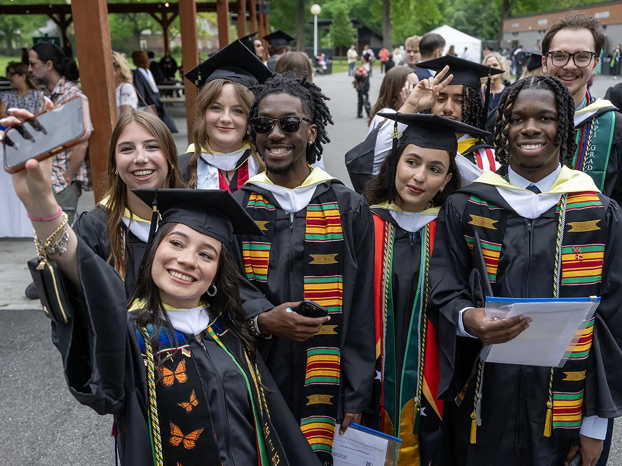 A group of joyful students in graduation caps and gowns gather closely to take a selfie. They are smiling and wearing stoles that represent diverse cultures and academic achievements, surrounded by fellow graduates and families in the background.