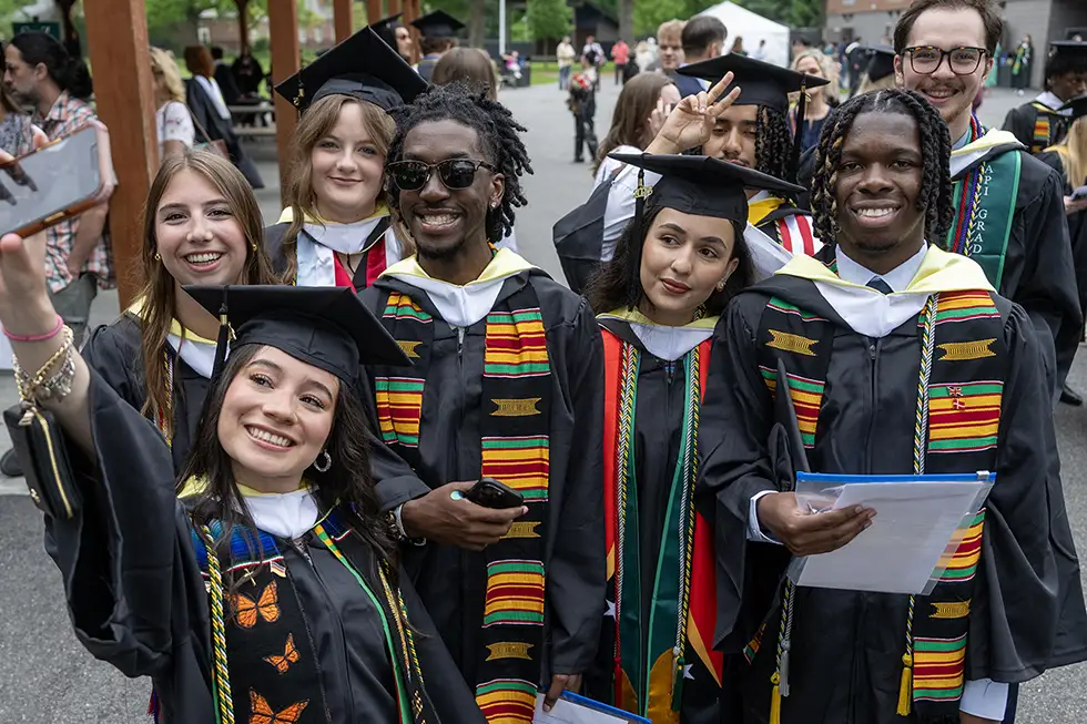 A group of diverse Skidmore College students in graduation caps and gowns pose for a joyful selfie, some wearing colorful stoles representing cultural heritage and academic honors.