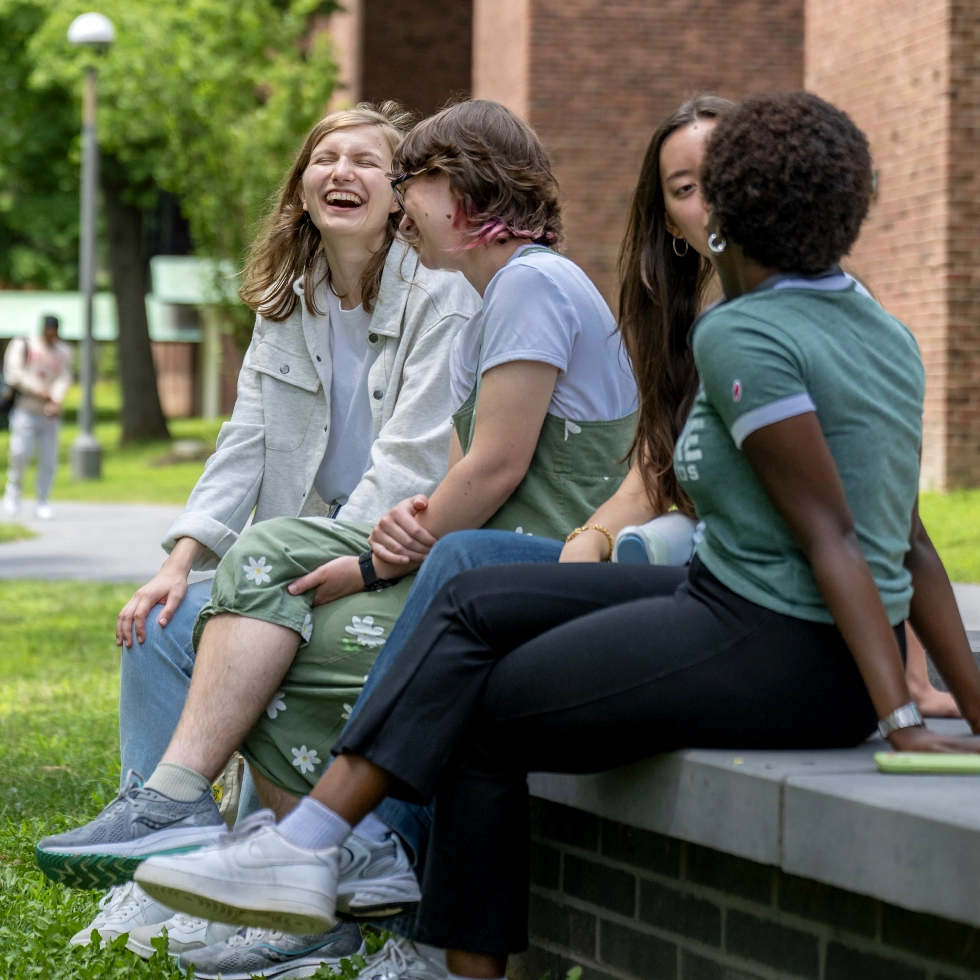A group of Skidmore students laughing and talking together on a sunny day outside a campus building.