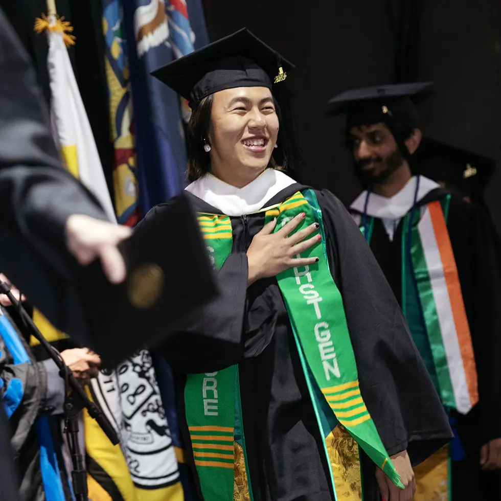 A smiling Skidmore College graduate stands with one hand over their heart during a commencement ceremony. The student wears a black cap and gown, a white academic hood, and a green Kente-style stole that reads “FIRST GEN” and “SKIDMORE.”