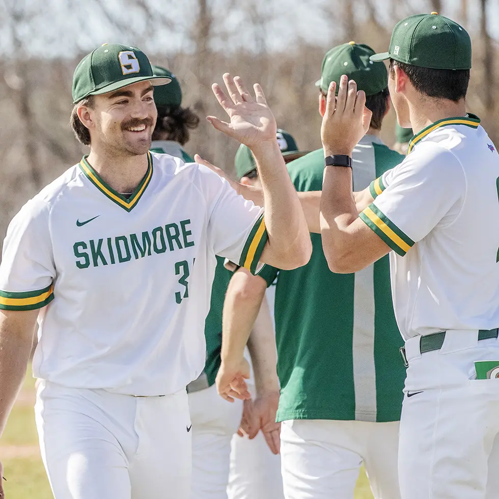 Skidmore College baseball players exchange high-fives on the field during a game or practice. The players wear white uniforms with green and gold trim and green caps featuring a bold "S" logo.