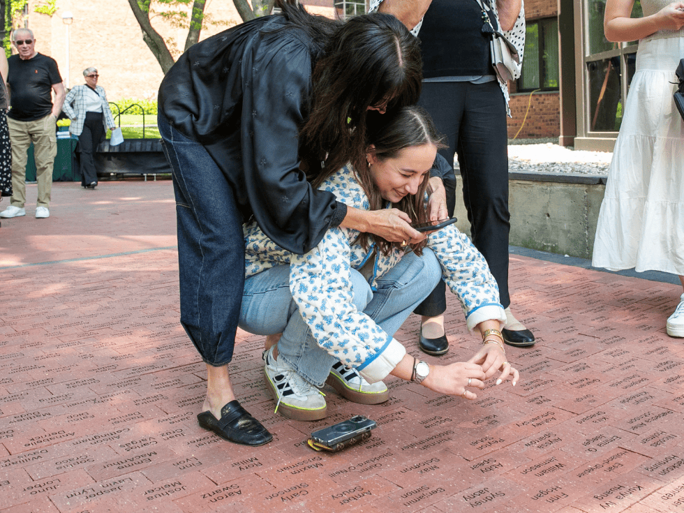 A student bends down to make a heart with her hands around her brick with her name on it, while her mother stands over her to take a photo.