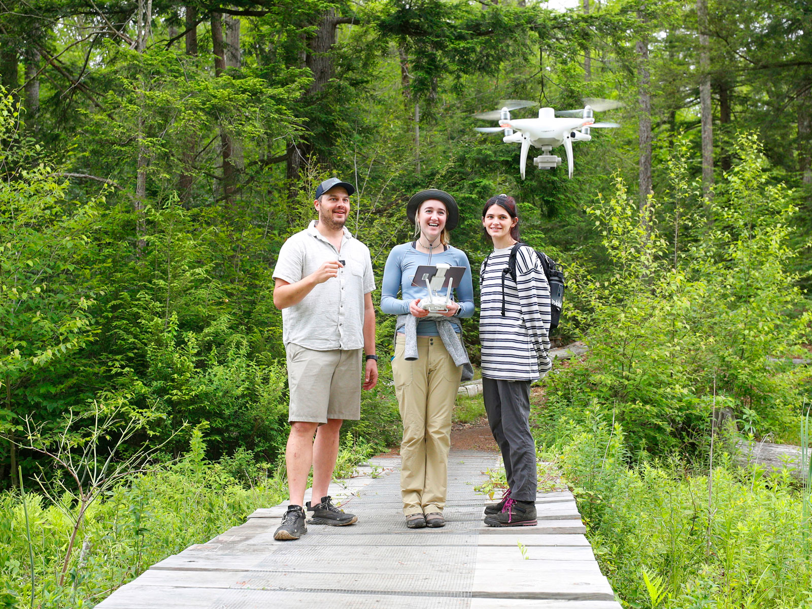 Three people standing on a wooden path in a forest, operating a drone with a remote control and tablet, engaged in a field research or GIS mapping activity.