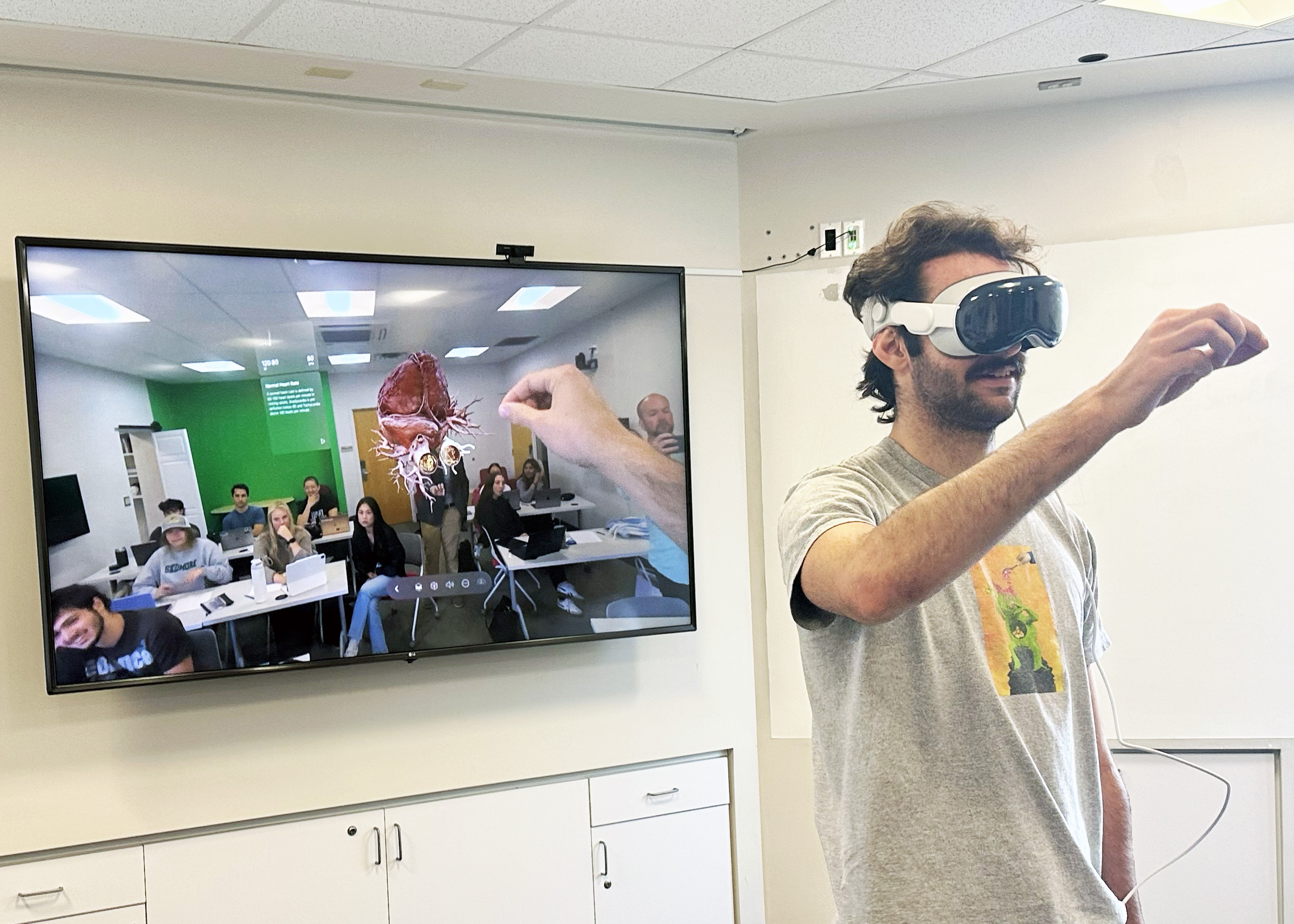 A student uses the Apple Vision Pro headset to manipulate the anatomically accurate human heart displayed on the screen behind them