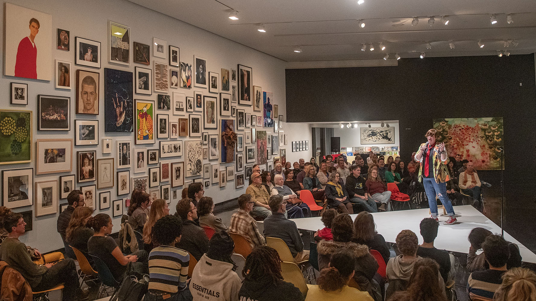 Beck stands on a triangular stage during their "Queerly Beloved" performance, a gallery wall can be seen to the left