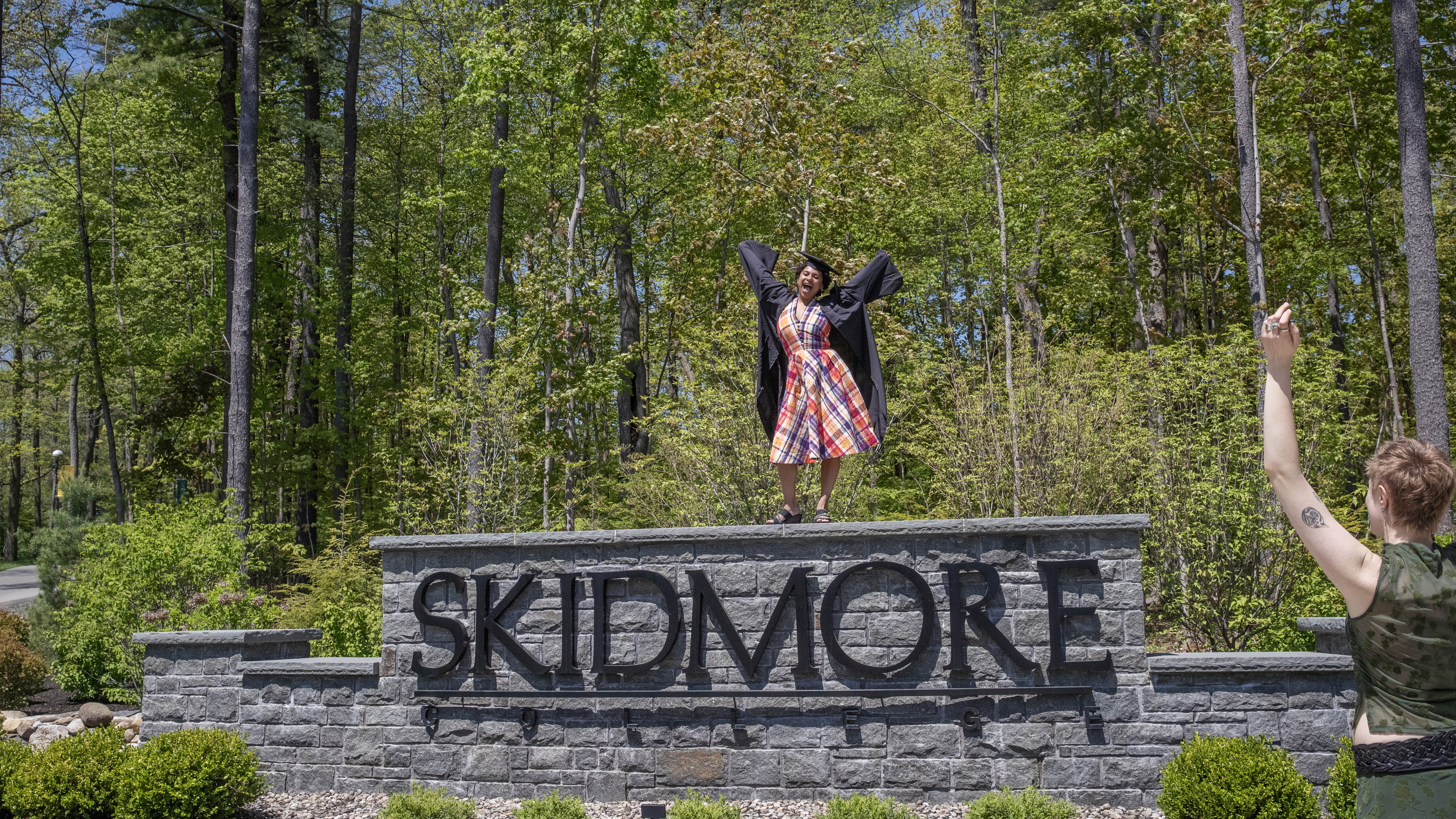 A senior celebrating on top of Skidmore's campus sign 