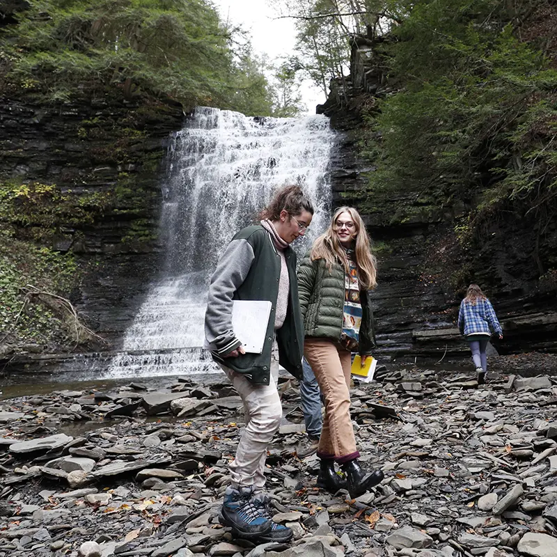 Skidmore students explore the rocky base of a scenic waterfall on a field trip, holding notebooks and smiling during a hands-on environmental science outing in upstate New York.