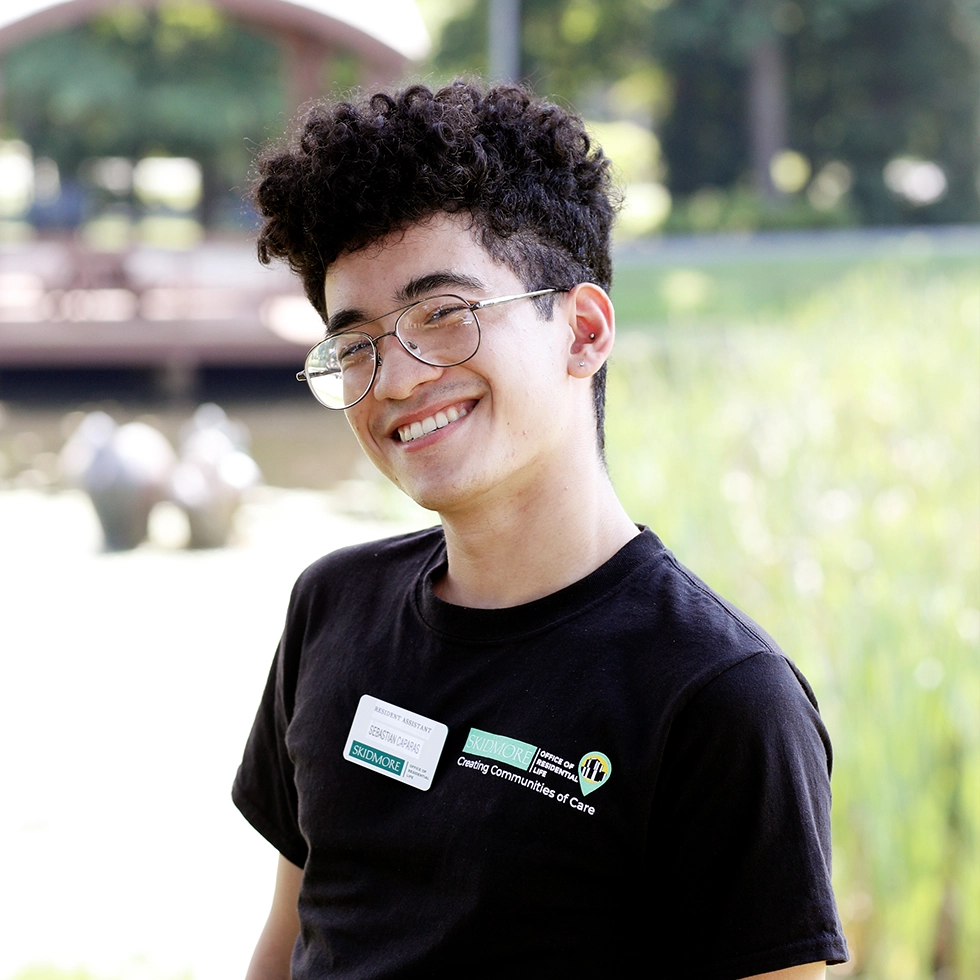 Skidmore resident assistant smiling outdoors near Haupt Pond, wearing a name tag and “Creating Communities of Care” T-shirt.