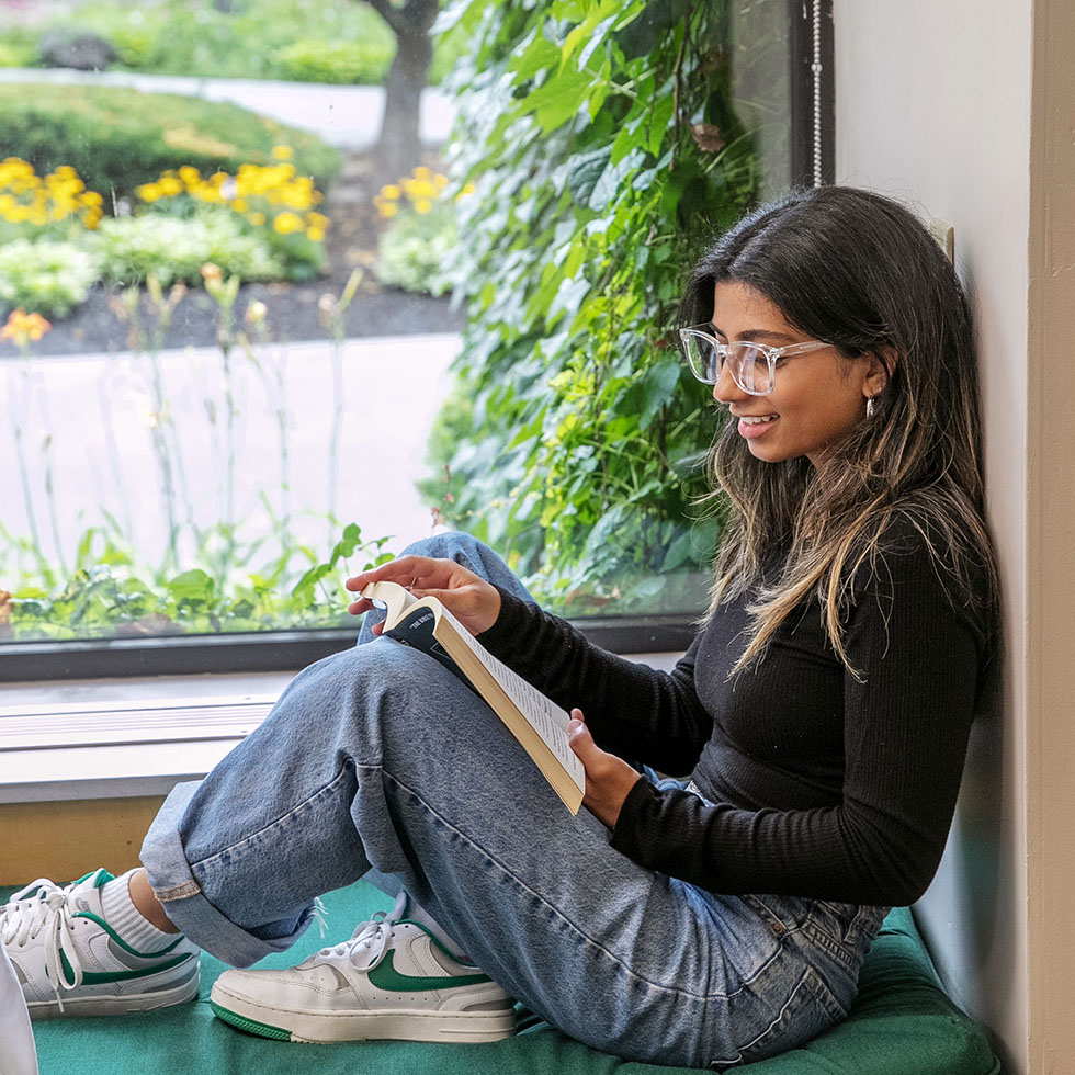 Skidmore student sitting by a large window seat with green ivy outside, reading a book and enjoying a quiet moment their dorm room.