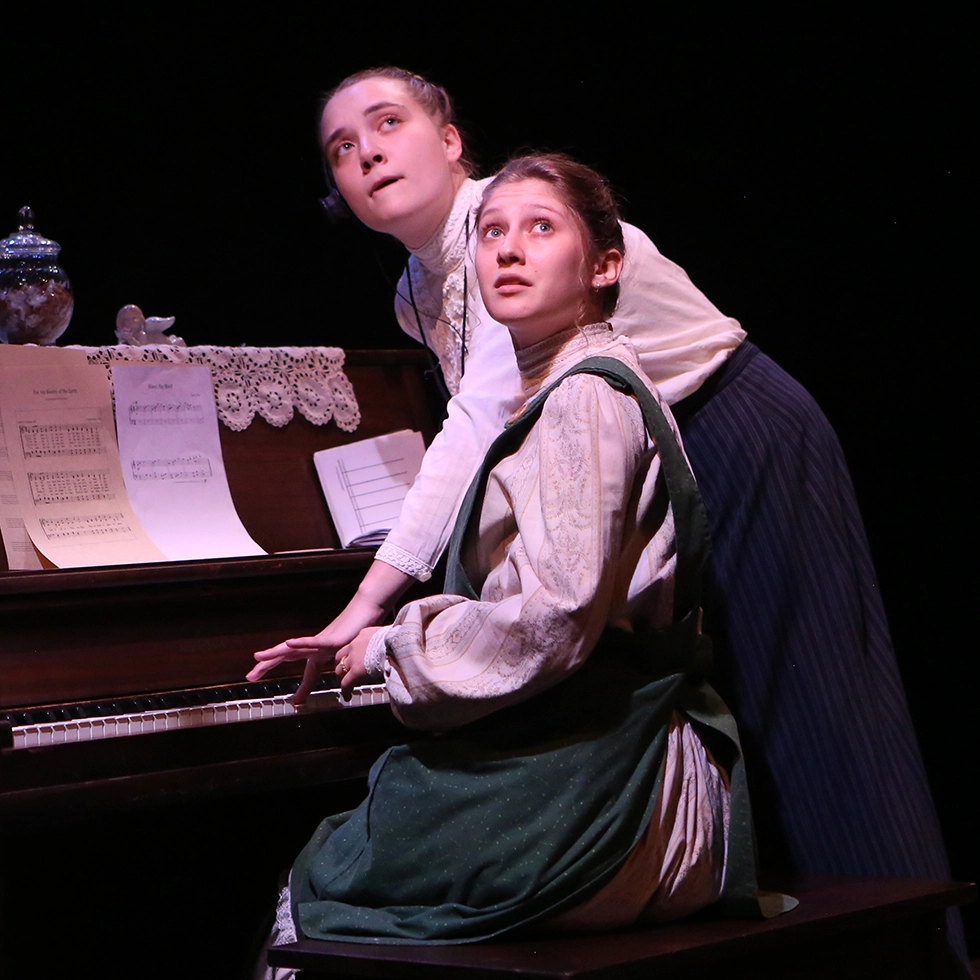 Two Skidmore students in period costume perform a dramatic scene at a piano during a campus theater production.