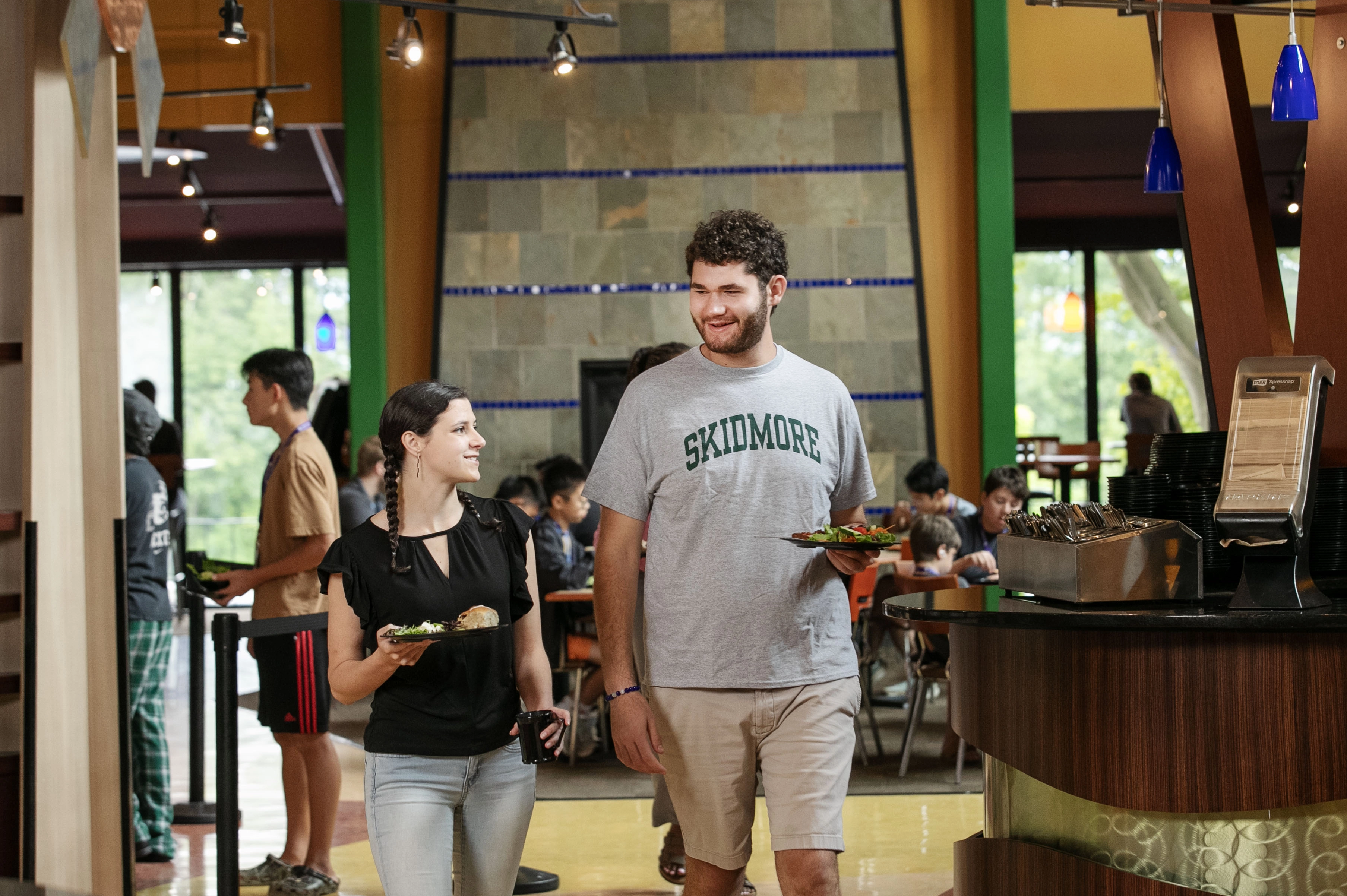 Two Skidmore students chatting and carrying plates of food in the college dining hall during a meal.