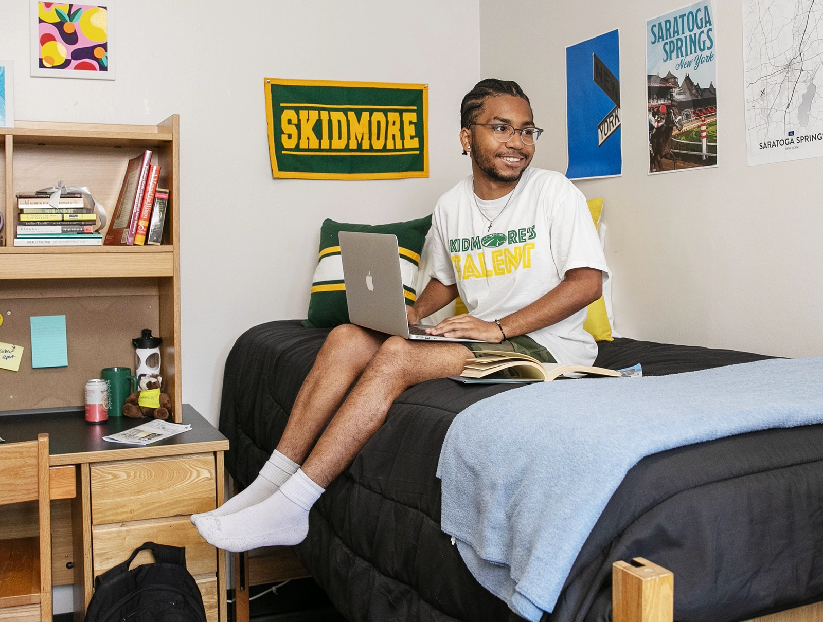 Skidmore student smiling while working on a laptop in a neatly decorated dorm room, surrounded by posters and personal touches.