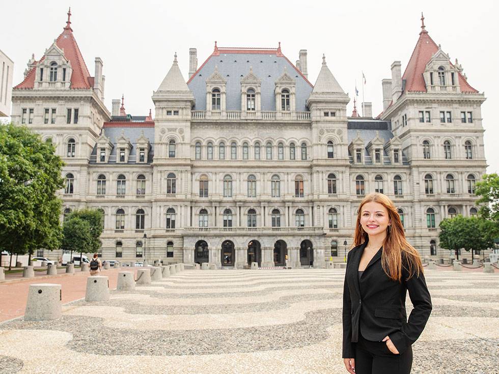 Cassidy Goda '28 stands outside the New York State Capitol building, wearing a black suit, smiling at the camera with one hand in her pocket.
