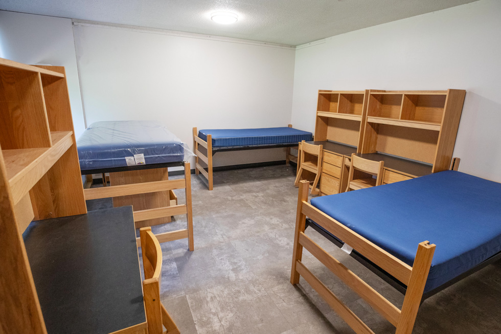 Triple residence hall room with blue mattresses, desks, shelves, and light wood furniture.