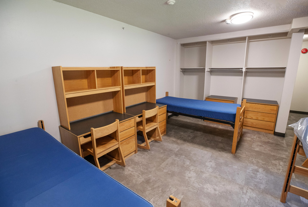 Triple residence hall room with blue mattresses, desks, shelves, and light wood furniture.