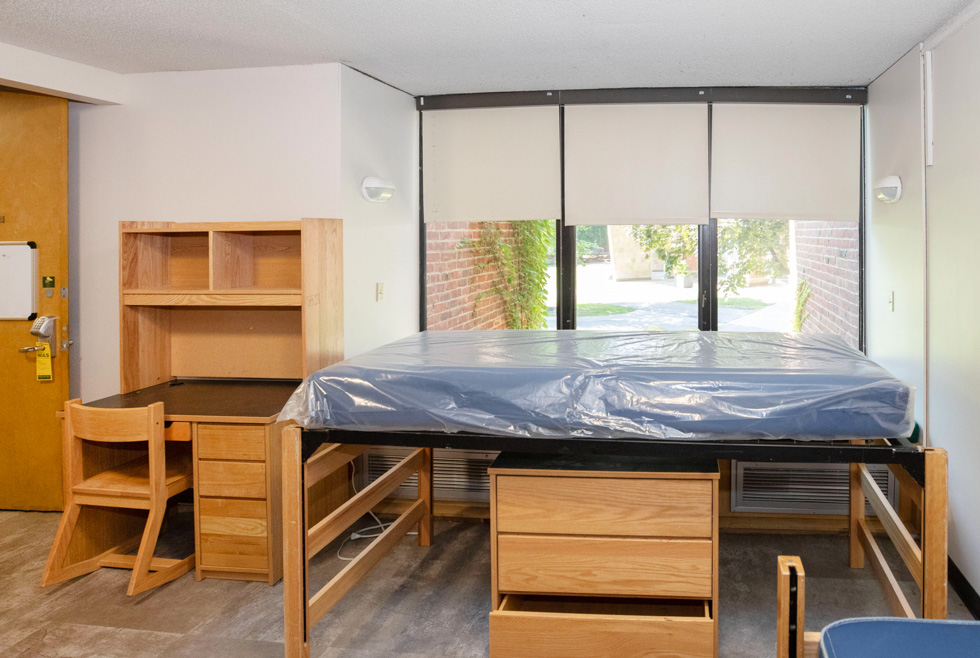 Triple residence hall room with blue mattresses, desks, shelves, and light wood furniture.