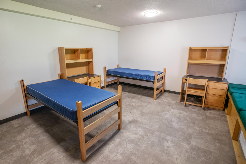 Double residence hall room with blue mattresses, desks, shelves, and light wood furniture.