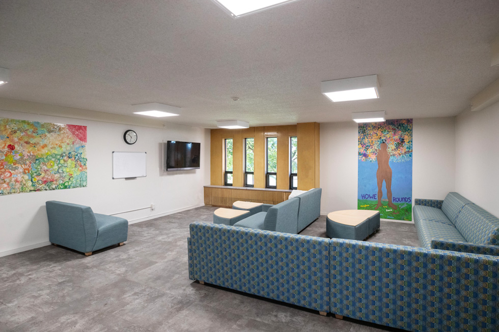 Residence hall lounge with blue couches, wall art, a mounted TV, and large windows.