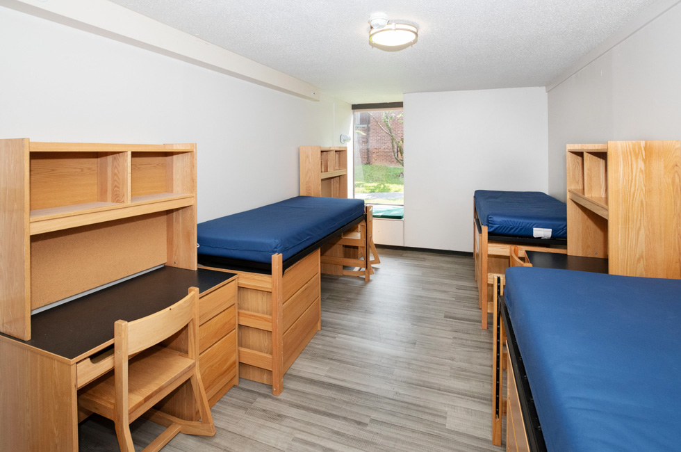 Triple residence hall room with three blue mattresses, desks, shelves, wood furniture, and a window seat.