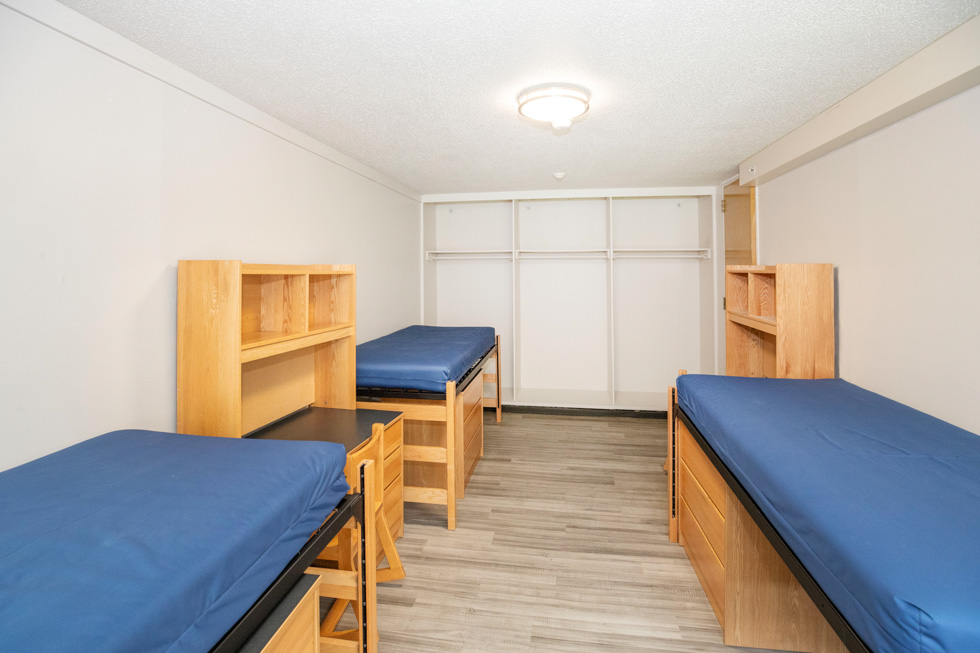 Triple residence hall room with three blue mattresses, desks, shelves, wood furniture, and a window seat.
