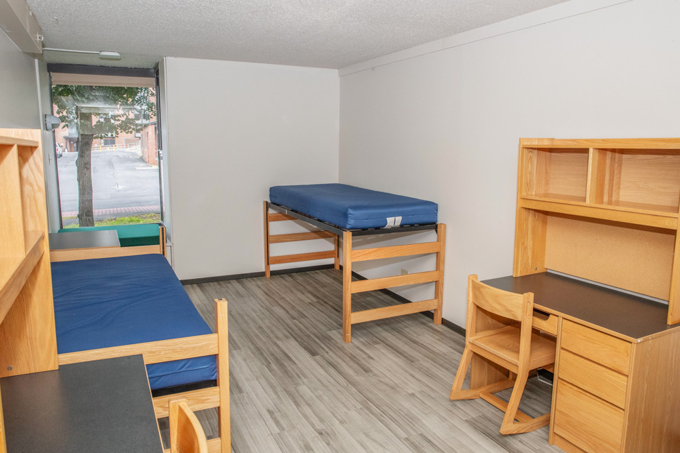 Double residence hall room with three blue mattresses, desks, shelves, wood furniture, and a window seat.