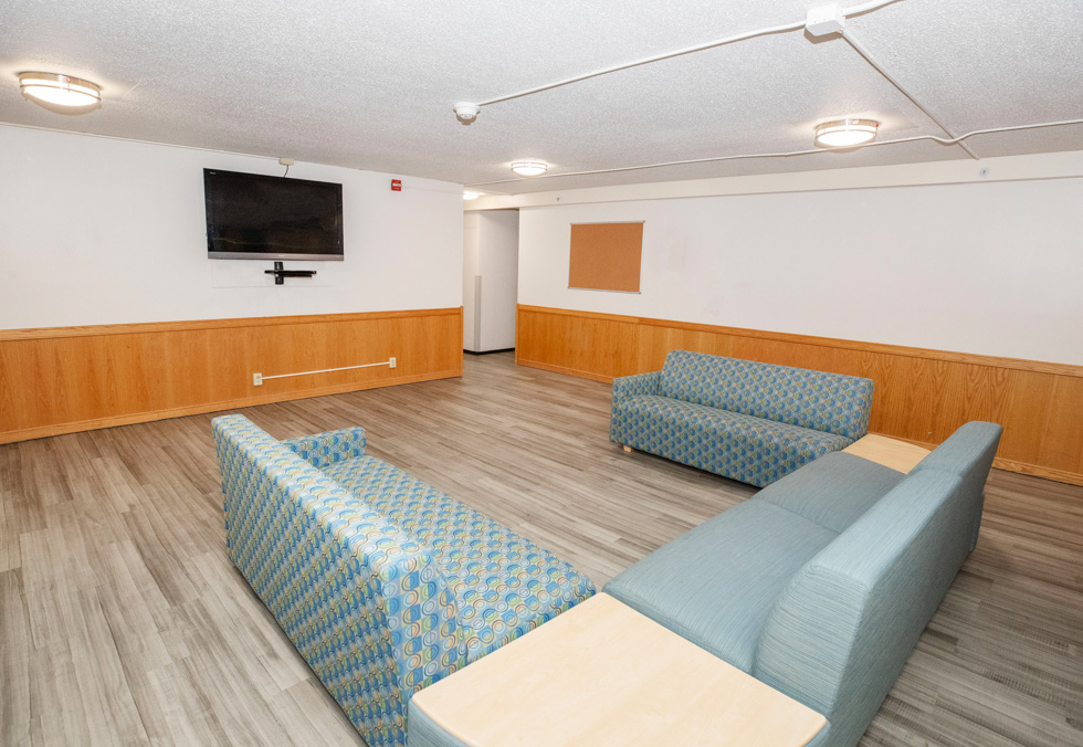 Residence hall lounge with blue couches, a wall-mounted TV, wood paneling, and open floor space.
