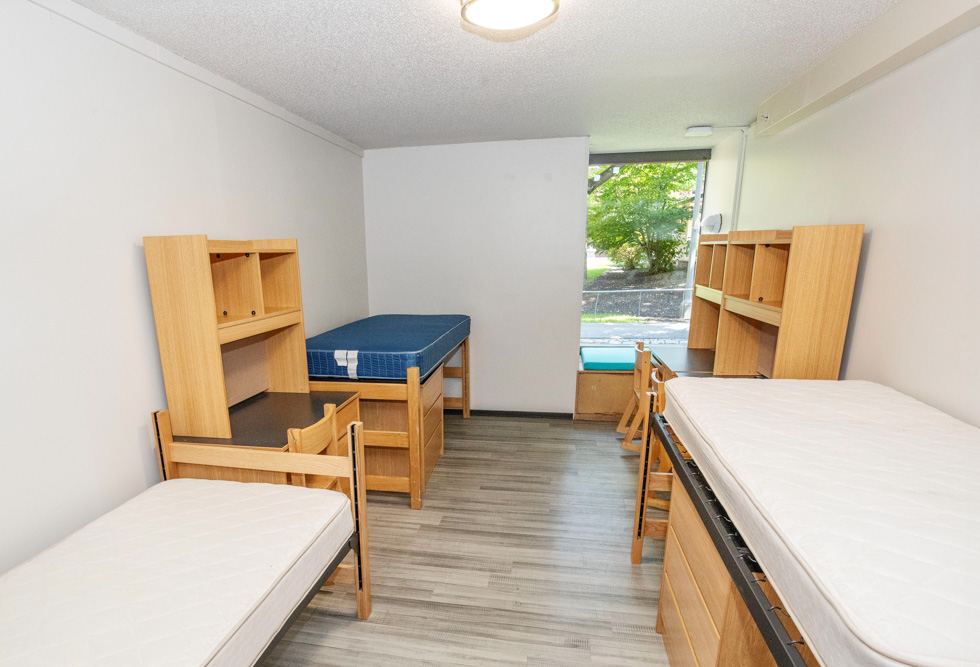 Empty McClellan residence hall room with three beds, desks, shelves, wood furniture, and a cushioned window seat.