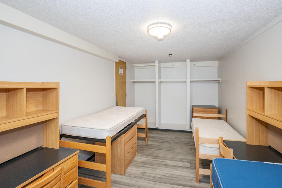 Empty residence hall room with three beds, desks, open closet space, and light wood furniture.