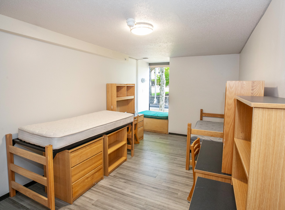 Empty residence hall double room with beds, desks, shelving, and open closet space.