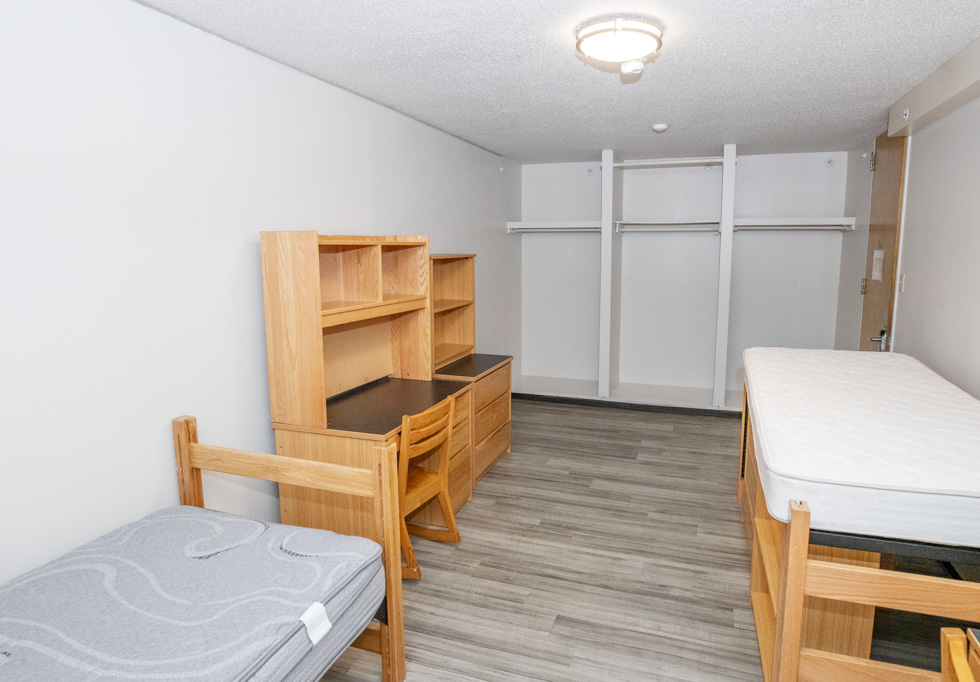 Empty residence hall double room with beds, desks, shelving, and open closet space.